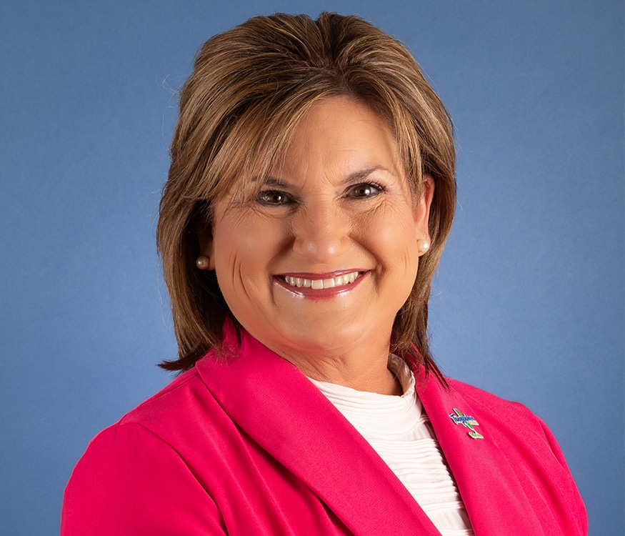 Connie Henry, a smiling woman with short brown hair, wears a bright pink blazer over a white top, pearl earrings, and a pin on her lapel as she stands in front of a plain blue background. She has a “Tangipahoa Parish” pin on his lapel and represents Tangipahoa Parish Government.