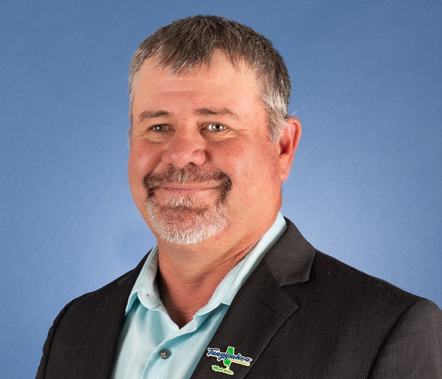Donnie Colona, a middle-aged man with short gray hair and a beard, is smiling in front of a plain blue background, wearing a suit jacket over a light blue shirt. He has a “Tangipahoa Parish” pin on his lapel and represents Tangipahoa Parish Government.