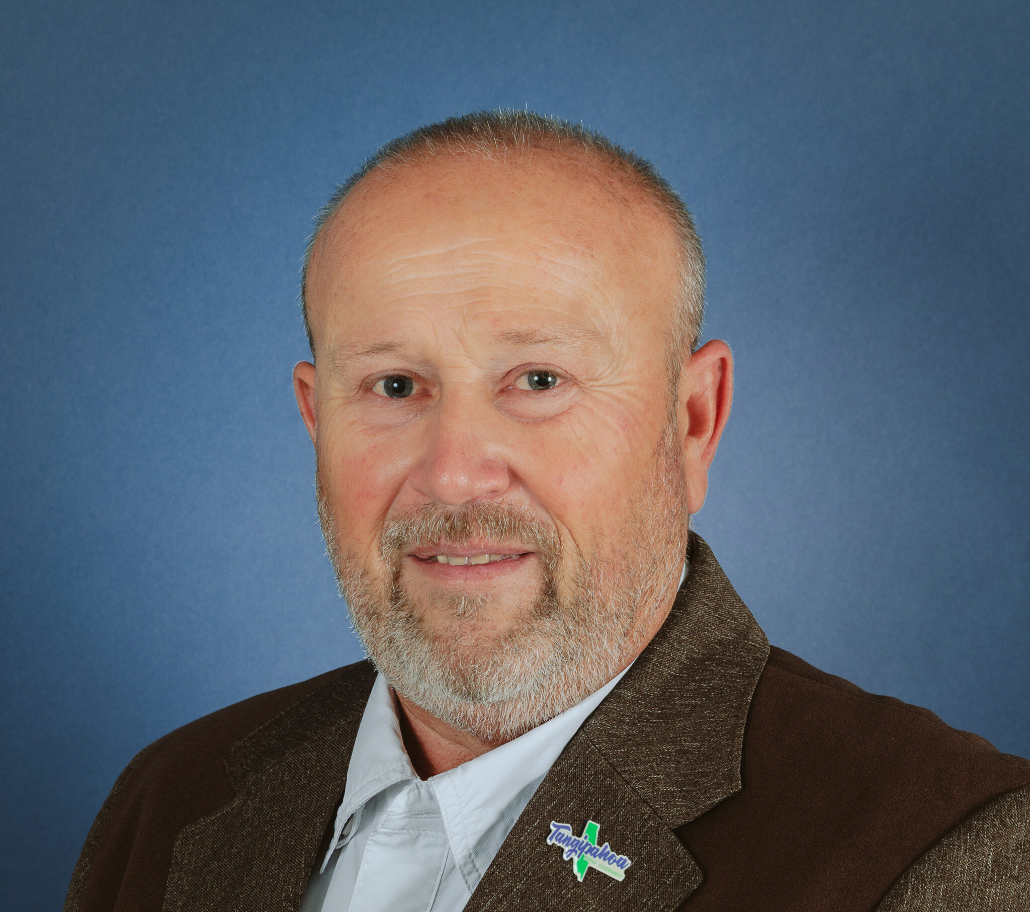 A middle-aged man with a light beard and short hair is wearing a brown jacket with a white collared shirt. He is smiling slightly and poses against a plain blue background.