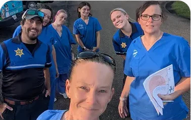 A group of seven people in blue uniforms, some with star-shaped badges, smile for a selfie outdoors. One person in front holds sunglasses, while another holds papers. The mood is friendly and casual.