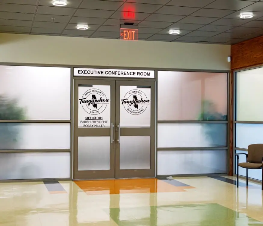 Entrance to an executive conference room with glass doors displaying logos for the Tangipahoa Parish Presidents Office. The hallway has colorful tile flooring and a chair against the wall.