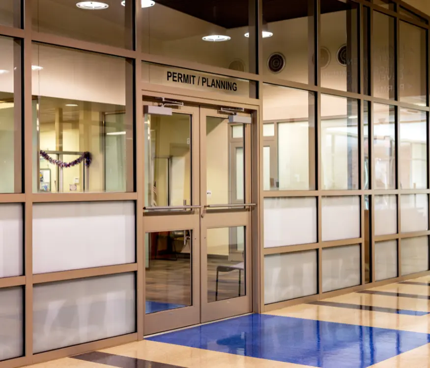 A glass-walled office with double doors labeled “Permit/Planning.” The interior appears empty, and the hallway outside has a blue and tan tiled floor.
