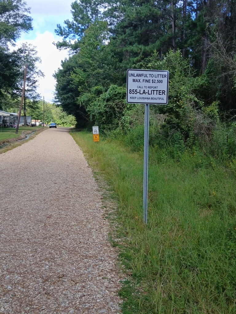 A gravel road with grass and trees on one side. A sign reads: Unlawful to litter. Max fine $2,500. Call to report 855-LA-LITTER. Keep Louisiana Beautiful. Another small sign is further down the road.