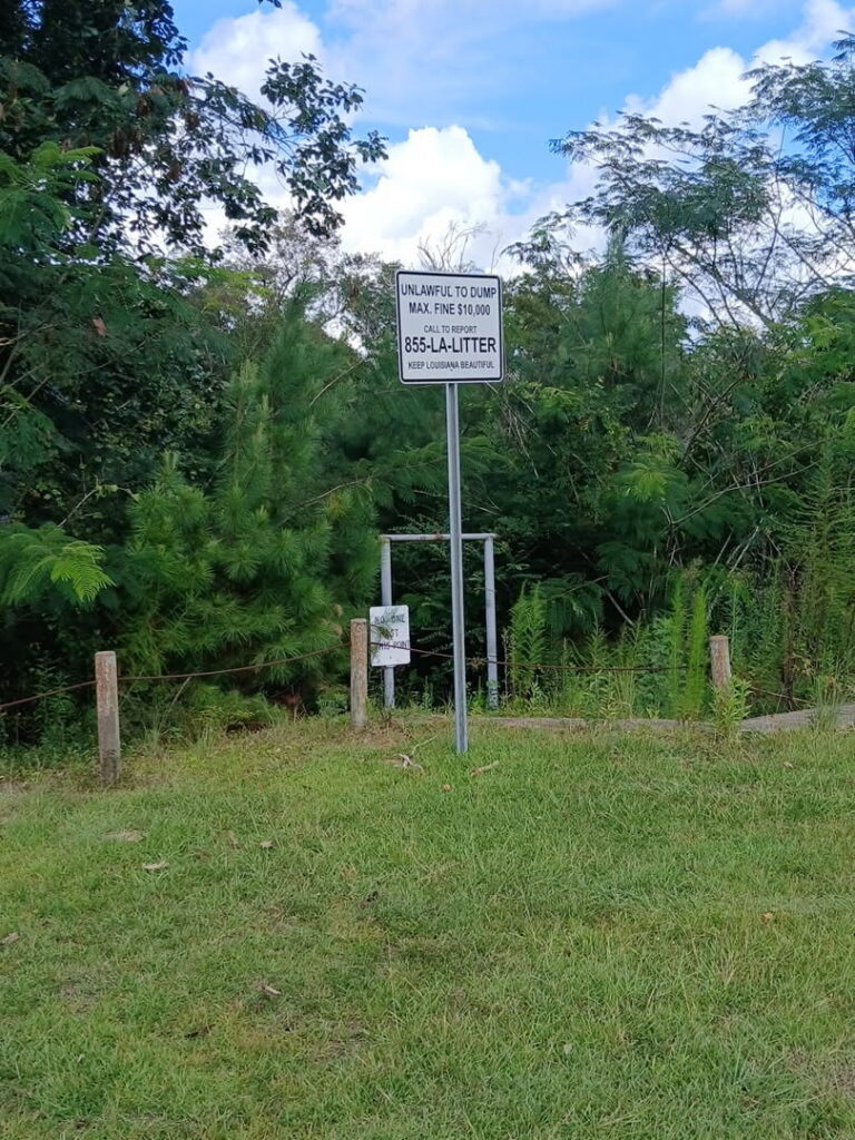 A sign on grass reads: Unlawful to dump. Max. fine $10,000. 855-LA-LITTER. Keep Louisiana beautiful. Trees and bushes fill the background under a partly cloudy sky.